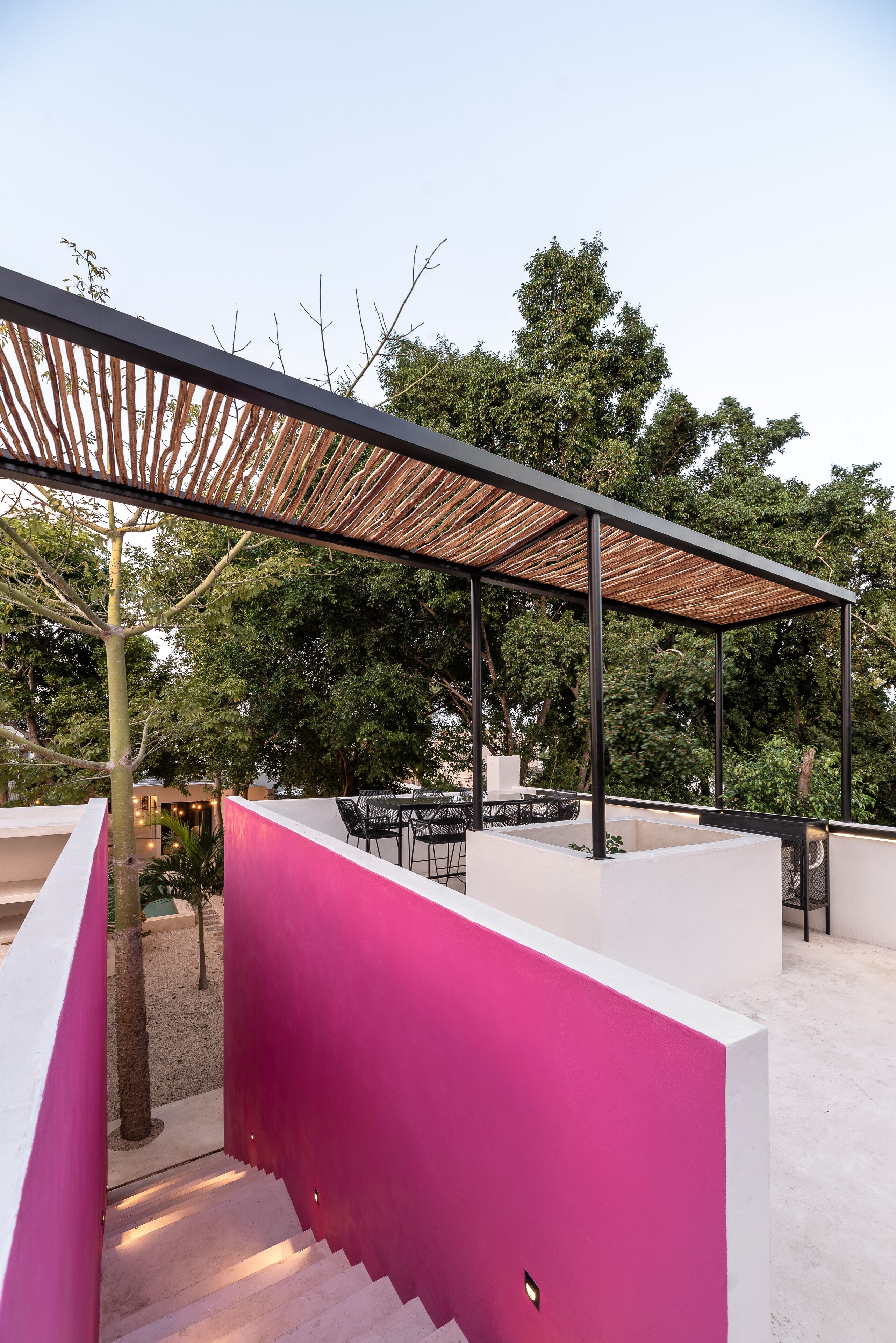Un patio exterior moderno con una pérgola de madera, paredes blancas y rosas, y un área de estar con mesas y sillas.