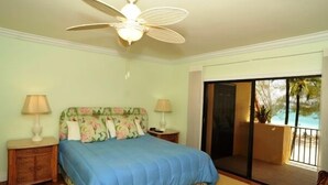 Un dormitorio con cama, dos lámparas de noche, ventilador de techo y vistas a un balcón con piscina.