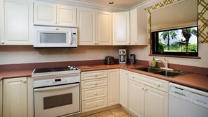 Una cocina con armarios blancos, microondas, estufa, cafetera y ventana con vista a palmeras.