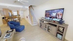 Ein geräumiges Wohnzimmer mit einem großen Flachbildfernseher, einem blauen Sessel und einer Holztreppe, die zu einer weiteren Ebene führt.