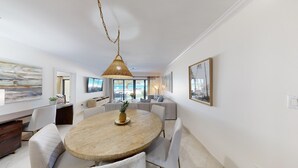 A modern dining area with a round wooden table, white upholstered chairs, and a hanging wicker chandelier. - Oceanfront Luxury, Spectacular 7 Mile Beach – Grd Floor|The Islands Club #24 (Grand Cayman)