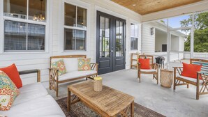 A covered patio with white walls, wooden ceiling, and large windows. There are wicker chairs, a wooden table, and a bench with colorful cushions.