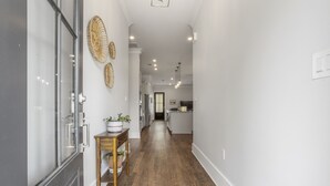 A hallway with wooden flooring, a glass door, a wooden table with potted plants, and a kitchen area in the distance.