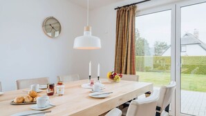 A dining area with a wooden table, white chairs, a hanging pendant light, a clock on the wall, and a view of a house through a glass door. - Zur Oie 27 "Westwind" (left) - Holiday home close to nature (Pruchten)