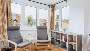 A modern room with two grey chairs, a small round table with a vase, and a bookshelf with books and decorative items. - Zur Oie 27 "Westwind" (left) - Holiday home close to nature (Pruchten)