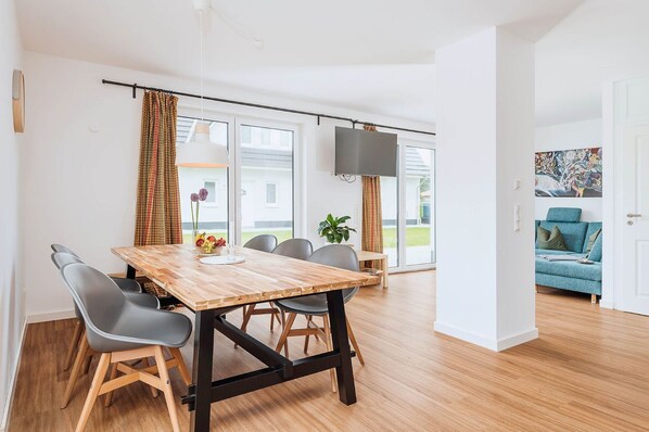 Un espace repas moderne avec une table et des chaises en bois, un téléviseur à écran plat et une vue sur l’extérieur grâce à de grandes fenêtres.