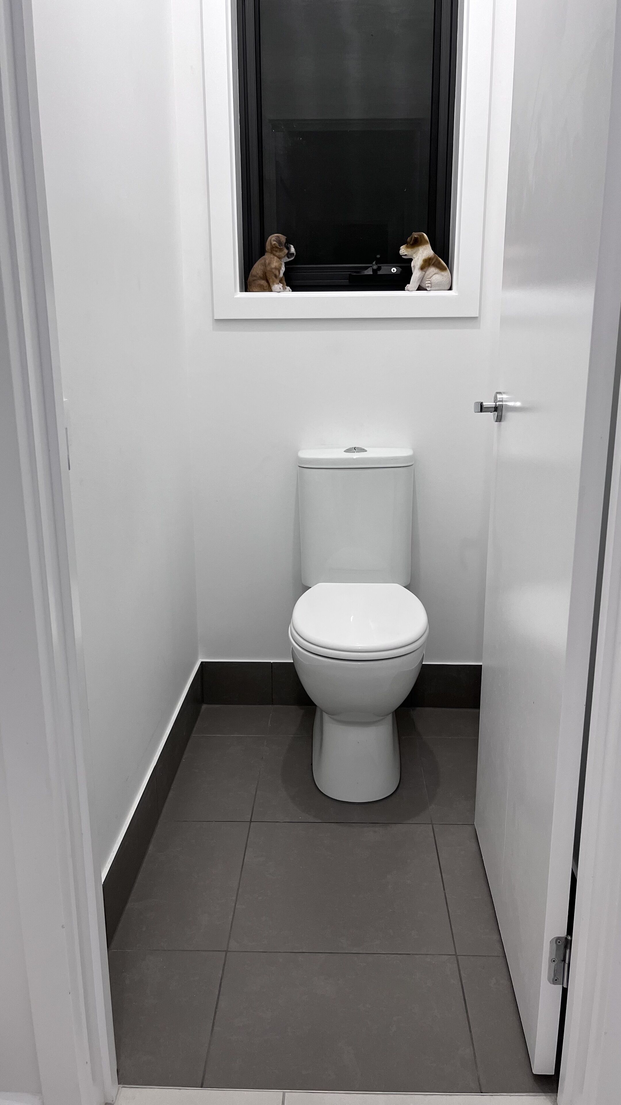 Une petite salle de bain blanche, avec des toilettes, une fenêtre située juste au-dessus et une porte sur le côté droit.