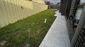 A residential backyard with a metal fence, a grassy lawn, and a concrete pathway leading to a brick building. - Modern House in Newton (Newton)