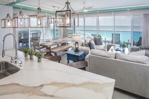 Et moderne køkken med marmor bordplade, et spiseområde med et træbord og stole, en grå sofa og udsigt til havet.