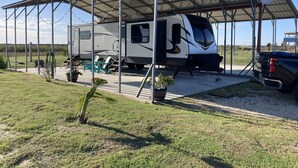 A recreational vehicle (RV) parked under a covered pavilion with a metal roof, surrounded by a grassy area and a small garden. - 10 minutes walk to the beach .Covered rv spot on concrete slab. NEW CAMPER (Bolivar Peninsula)