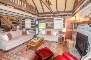 Interior de cabana de tronco com dois sofás, uma poltrona de couro vermelha, uma mesa de centro de madeira, uma lareira e uma escada.