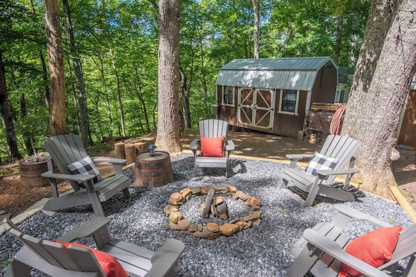 Uma área de estar rústica ao ar livre, com cadeiras Adirondack e um poço de fogo cercado por pedras, em meio a uma área arborizada com uma cabana de madeira ao fundo.