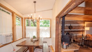 Ein gemütliches Blockhaus-Interieur mit einem Holzesstisch, Bänken, einer Pendelleuchte, einem Kamin und einem großen Fenster mit Vorhängen.