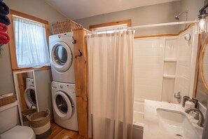 Ein kleiner Waschbereich mit Waschmaschine und Trockner, eine Dusche mit Holztür sowie ein Waschbecken mit Spiegel.