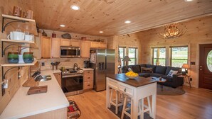 A cozy cabin interior with wooden walls and ceilings, a kitchen with modern appliances, a dining area, and a living room with a fireplace. - Jones River Cabin | Riverfront Stay with Hot Tub | Near West Jefferson (Creston)