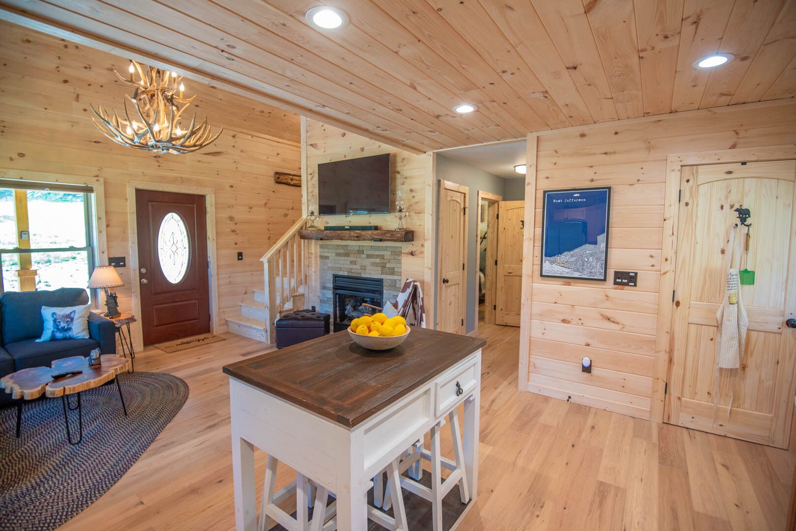 A cozy cabin interior with a fireplace, wooden walls, a dining area with a bowl of fruit, and a staircase leading to another room.