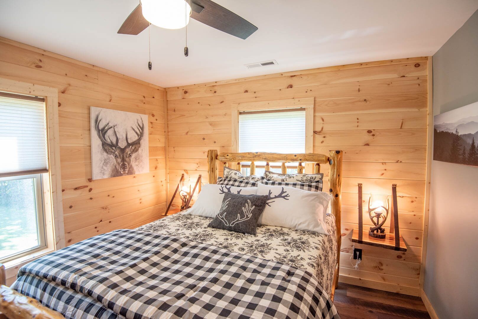 Une chambre à coucher dans un chalet en bois, avec un lit, une literie à carreaux, un ventilateur de plafond et un tableau représentant un cerf accroché au mur.