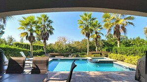 Zona de piscina con sillones de descanso y una mesa, rodeada de palmeras y un seto.