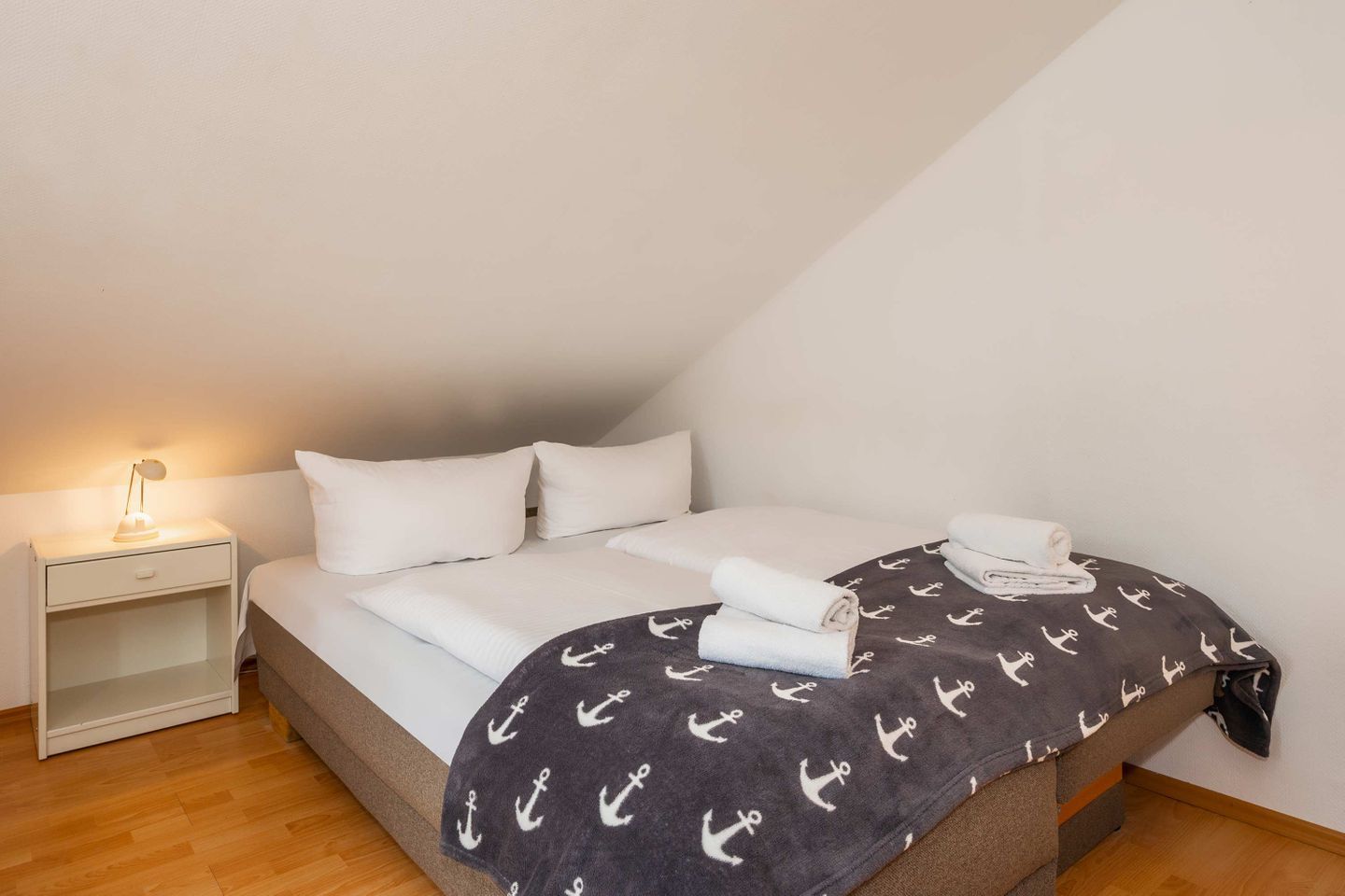 A single bed with white bedding, a dark blanket with anchor patterns, and a bedside lamp.