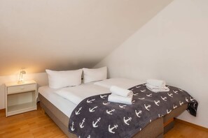 Una cama individual con ropa de cama blanca, un edredón oscuro con estampado de anclas y una lámpara de noche.