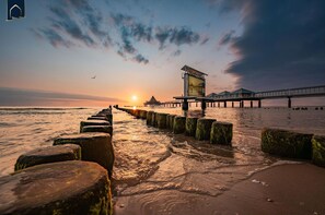 A pier extending into the water with a building at the end, a sunset in the background, and a row of rocks leading to the shore.