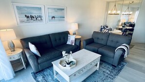 Ein modernes Wohnzimmer mit einem grauen Sofa, einem weißen Couchtisch und einem blauen Teppich mit Muster. An der Wand hängen zwei gerahmte Bilder, auf einem Beistelltisch steht eine Lampe und im Hintergrund ist ein Essbereich zu sehen.