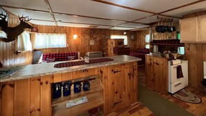 L’intérieur d’une cabane est aménagé avec du bois, il comprend un coin cuisine avec un poêle et une table à manger recouverte d’une nappe à carreaux.