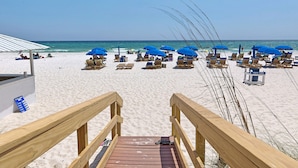 Eine Holztreppe führt zu einem Strand mit blauen Sonnenschirmen und Holzliegen.
