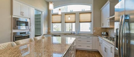 A modern kitchen with a large island, stainless steel appliances, and a view of the outdoors through large windows.