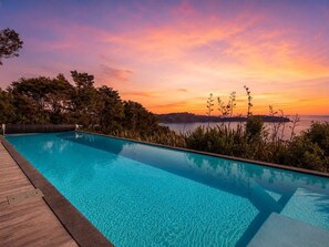 A clear swimming pool with a wooden deck, surrounded by greenery and overlooking a body of water with a sunset view.