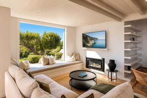 A modern living room with a sofa, a fireplace, a TV, and a view of greenery.