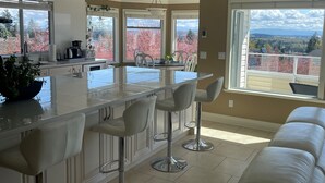 Una cocina moderna con una isla central, taburetes de bar y ventanas amplias que ofrecen vistas a árboles y un paisaje lejano.
