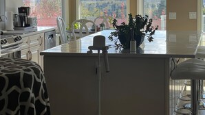Una cocina con una isla grande, sillas blancas, una cafetera y vistas a montañas a través de las ventanas.