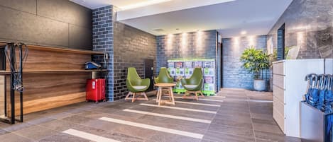 Un espace d’accueil avec un bureau en bois, des chaises vertes, une valise rouge et un distributeur automatique.