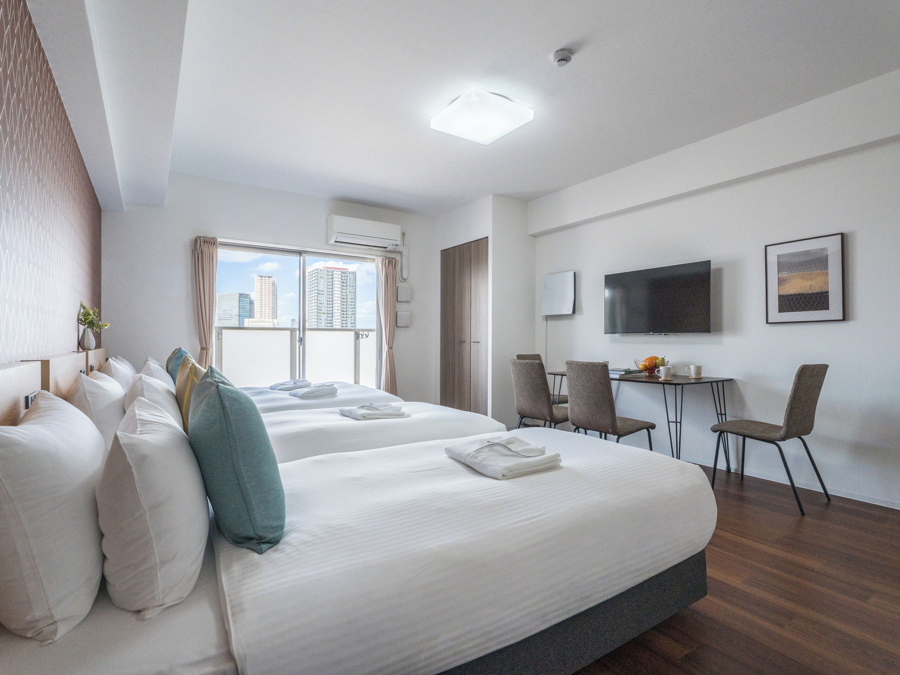 A hotel room with two beds, a desk with chairs, a flat-screen TV, and a window with a city view.