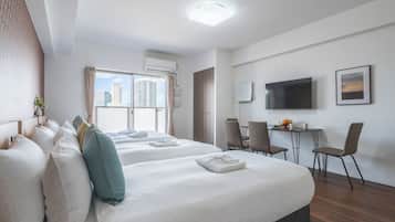 A hotel room with two beds, a desk with chairs, a flat-screen TV, and a window with a city view.