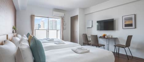 A hotel room with two beds, a desk with chairs, a flat-screen TV, and a window with a city view.