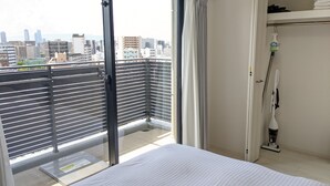 Quarto de hotel com uma grande porta de vidro deslizante para uma varanda, uma cama, uma estante com toalhas dobradas e um aspirador de pó.