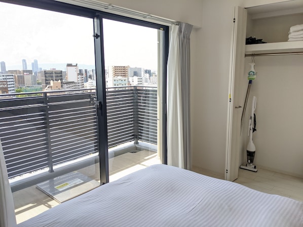 Habitación de hotel con un amplio ventanal corredero a un balcón, una cama, un estante con toallas dobladas y un aspirador.