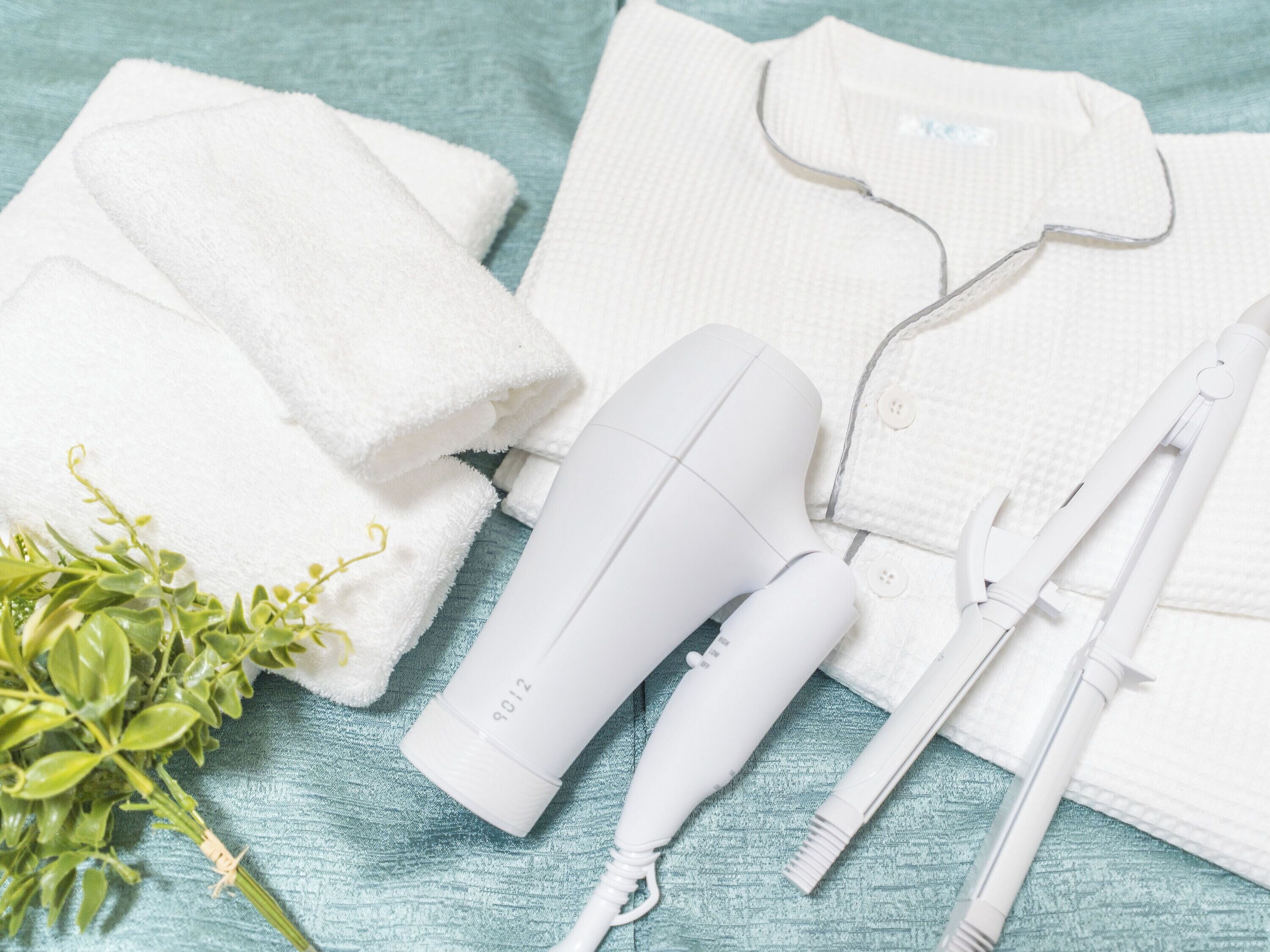 A white hair dryer, a white towel, a white shirt, and a pair of white scissors on a teal textured surface.
