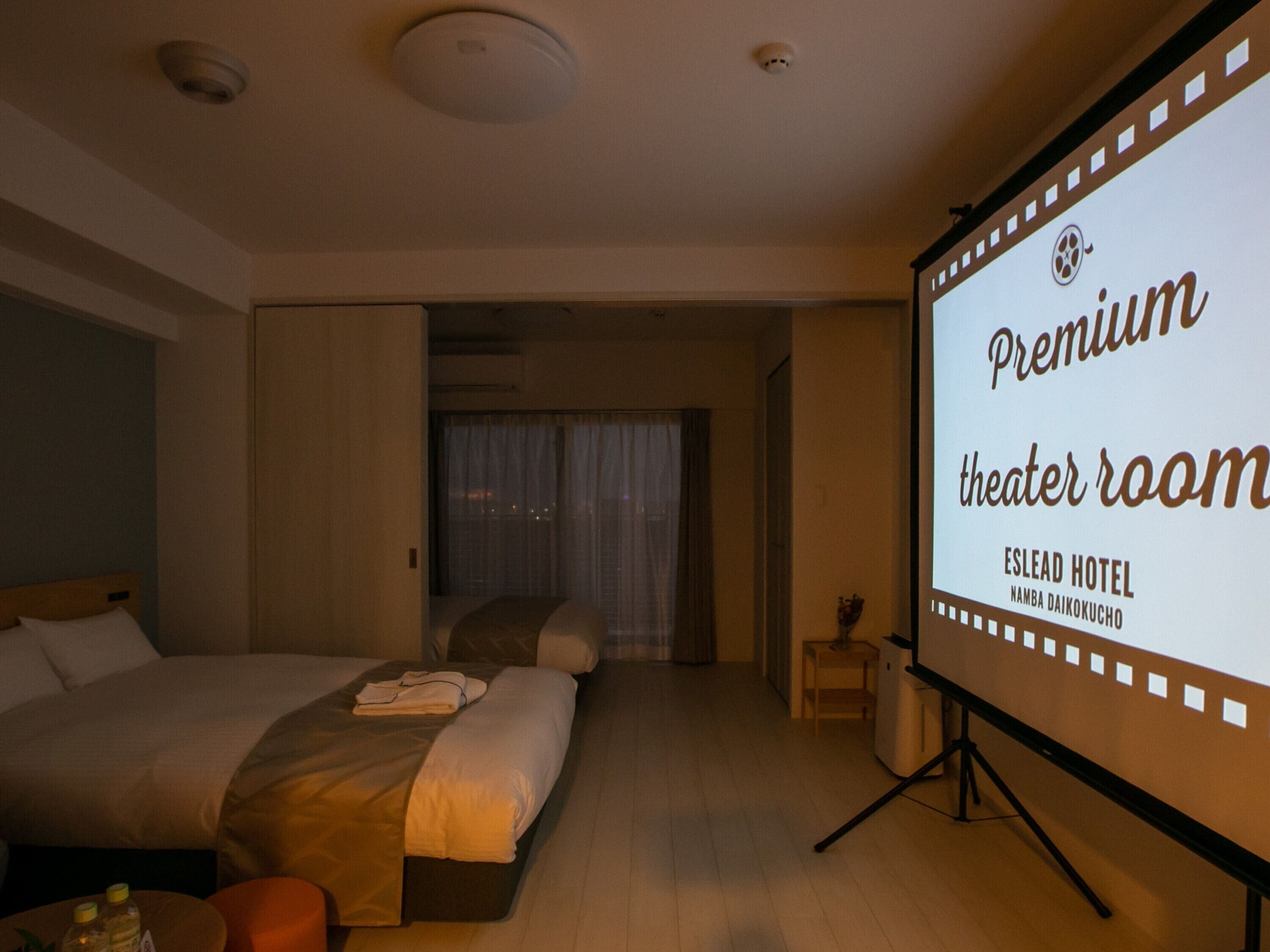 A hotel room with a large bed, a small table, and a projector screen displaying 'Premium theater room' text.
