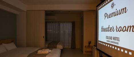 A hotel room with a large bed, a small table, and a projector screen displaying 'Premium theater room' text.