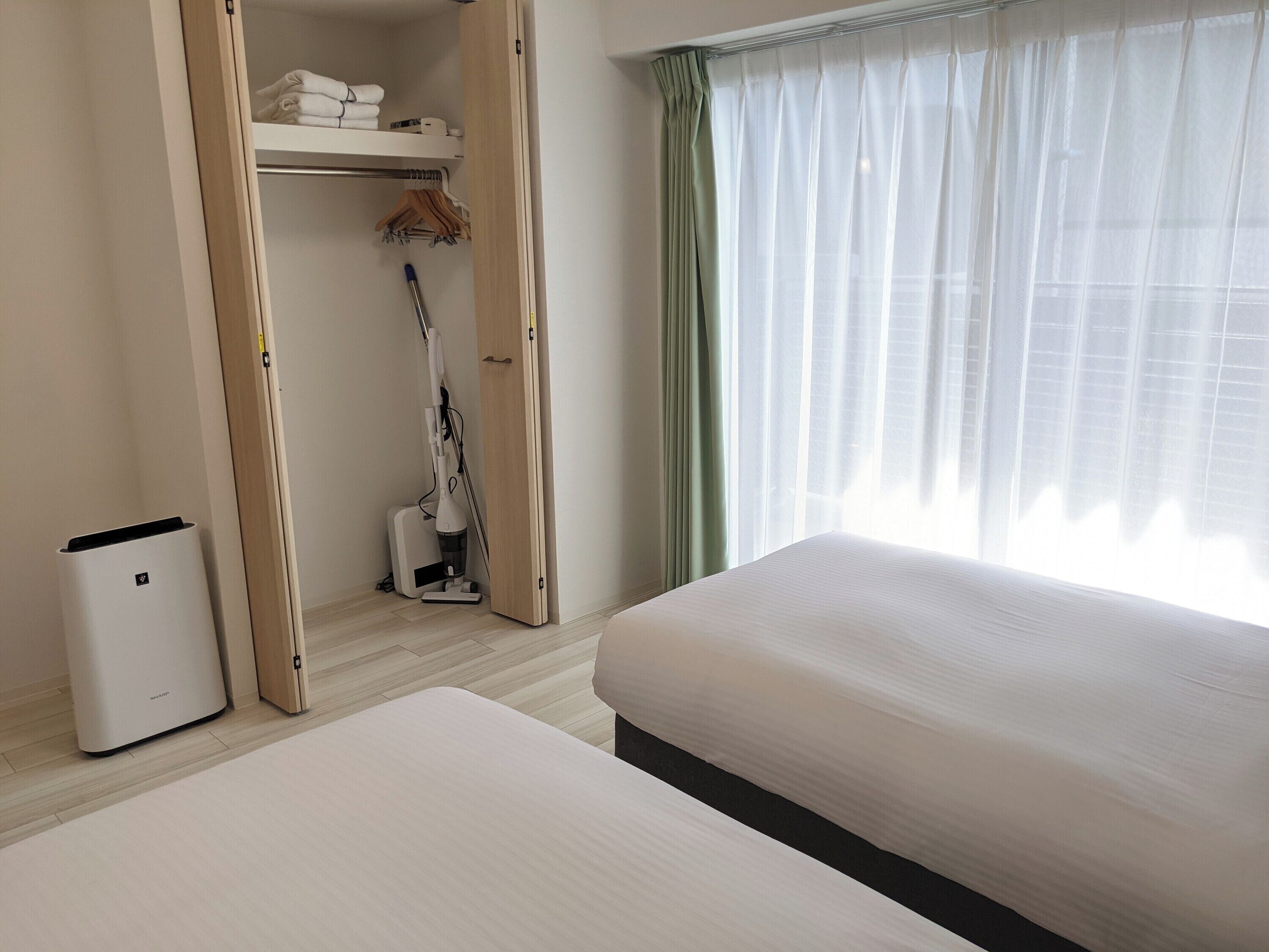 A hotel room with a bed, a closet with folded towels, a vacuum cleaner, and a window with sheer curtains.