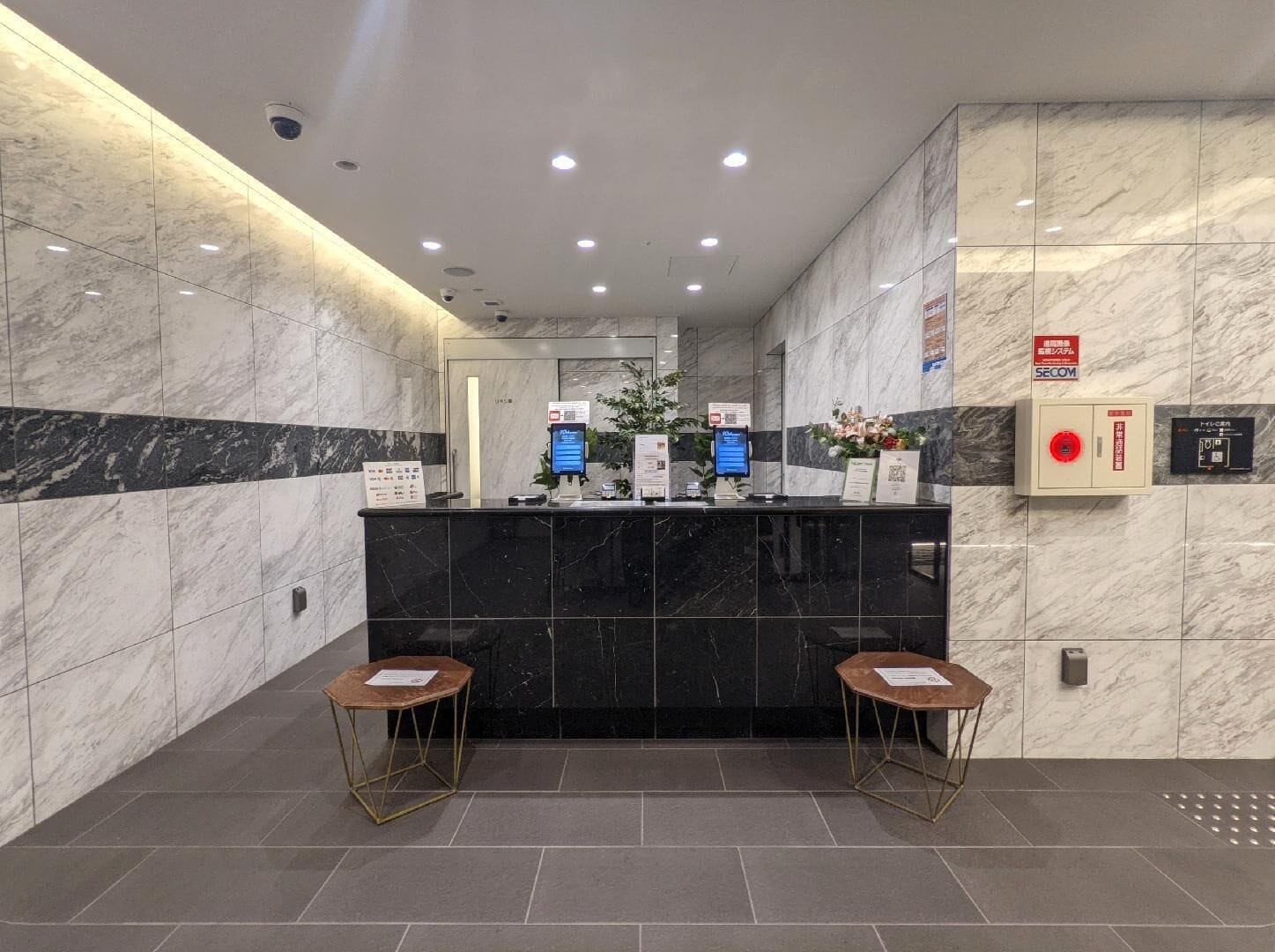 A reception area with a marble wall, a black reception desk, two wooden stools, and a fire alarm box.