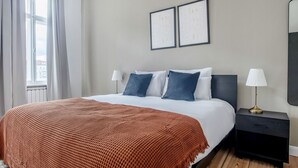 Une chambre d’hôtel avec un grand lit, deux tables de chevet, une commode, une fenêtre avec des rideaux et deux tableaux encadrés accrochés au mur.
