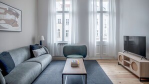 Un salon moderne avec un canapé gris, une petite table ronde, un téléviseur à écran plat et une fenêtre agrémentée de rideaux blancs.