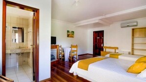 A hotel room with a wooden floor, a bed with white bedding and yellow pillows, a small table with chairs, a wooden wardrobe, and a bathroom visible through an open door. - Imperial Parque Hotel (Nova Petropolis)
