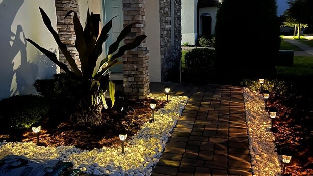 Een goed verlicht pad dat leidt naar een gebouw met stenen bekleding, een deur en ramen, omringd door planten en decoratieve verlichting.