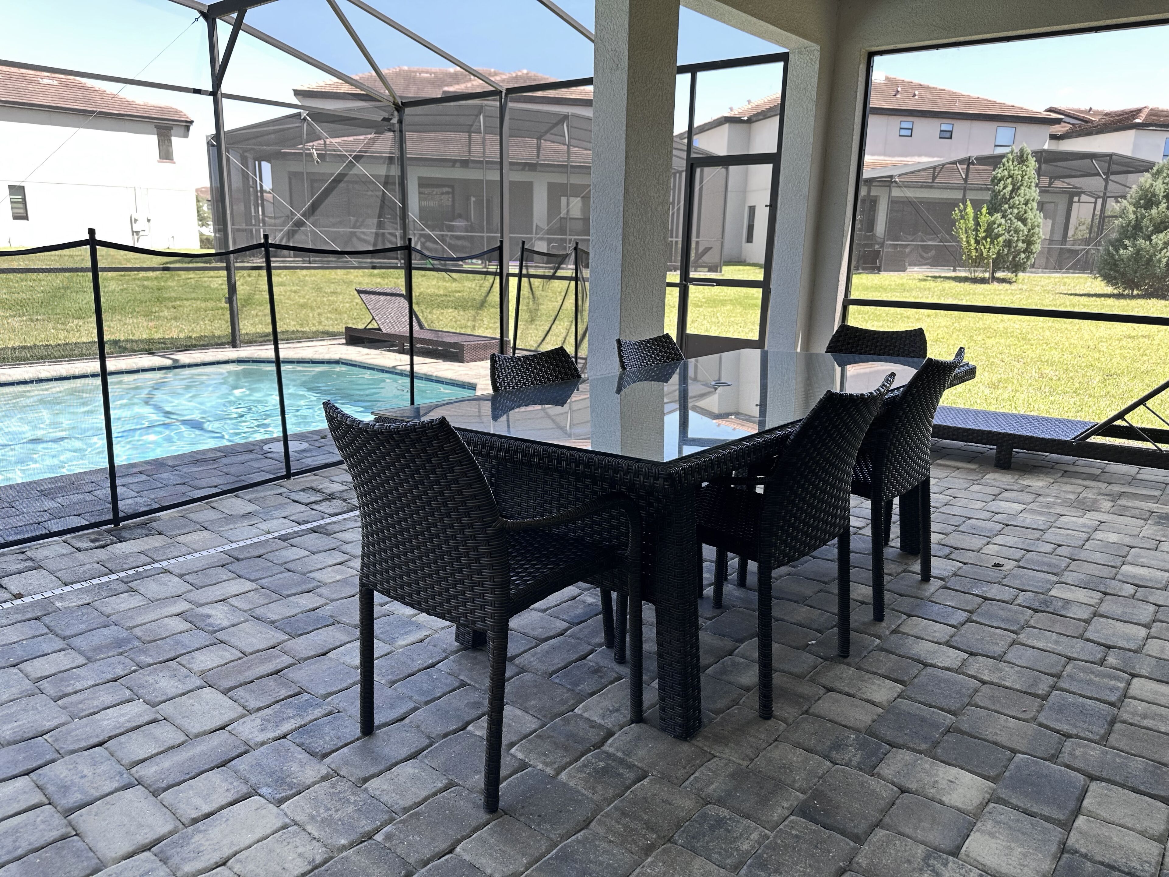 Une terrasse extérieure avec une table en verre et des chaises en osier, donnant sur une piscine et une pelouse, avec des maisons en arrière-plan.