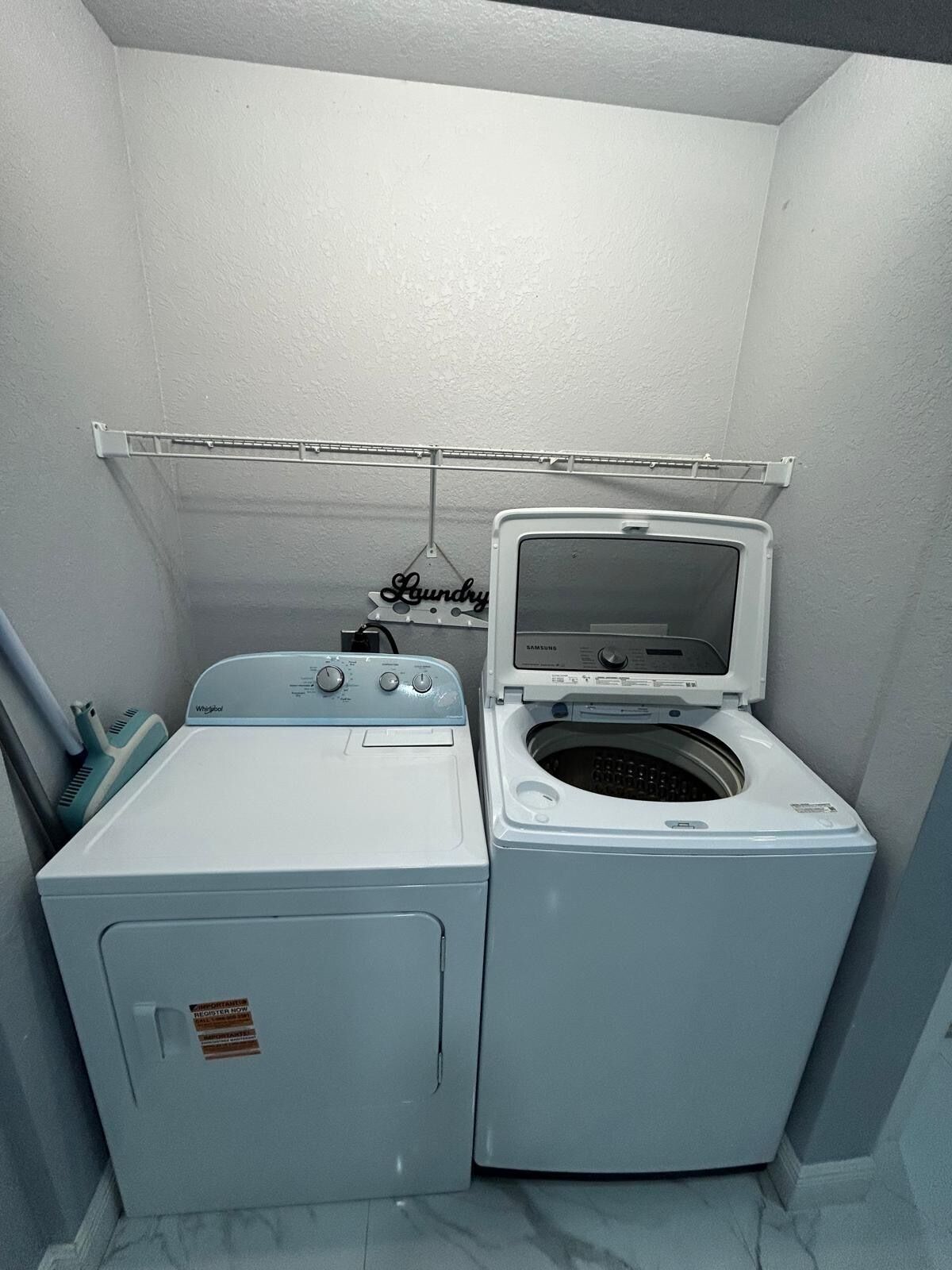 Une petite buanderie équipée d’un lave-linge et d’un sèche-linge à chargement frontal, d’un étendoir métallique et d’une enseigne fixée au mur.